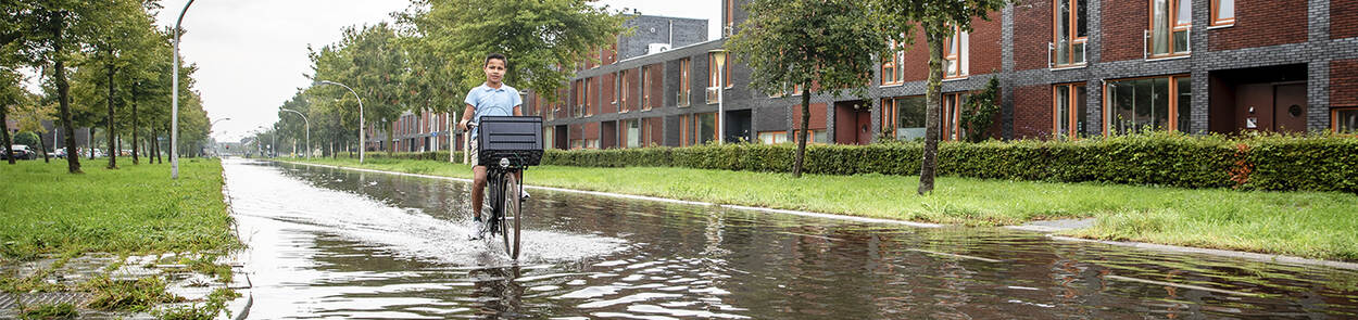 Een jongen fietst door een ondergelopen straat in een woonwijk na hevige regenval. De foto laat zien wat de impact is van extreme regen.