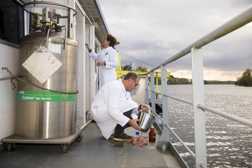 Twee medewerkers voeren werkzaamheden uit voor de Waterkwaliteit. De persoon voor in beeld schenkt natuurwater in een bruine fles.