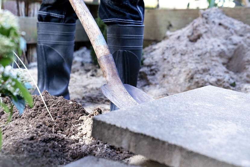 Een persoon met zwarte laarzen wipt een tegel uit de tuin met een schep om ruimte te maken voor groen en betere waterafvoer.