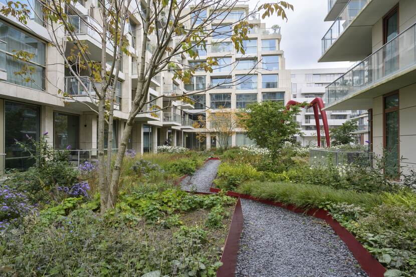 Een groene binnentuin met planten en bomen tussen appartementen. Er loopt een kiezelpad door de tuin met een rode rand als afscheiding.