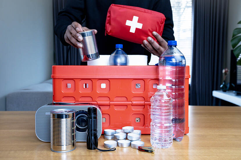 Een persoon stopt spullen voor een noodpakket in een rood krat. In het noodpakket zitten waterflessen, een EHBO-kit, een radio op batterijen, waxinelichtjes, batterijen, eten in blik en een zaklamp.