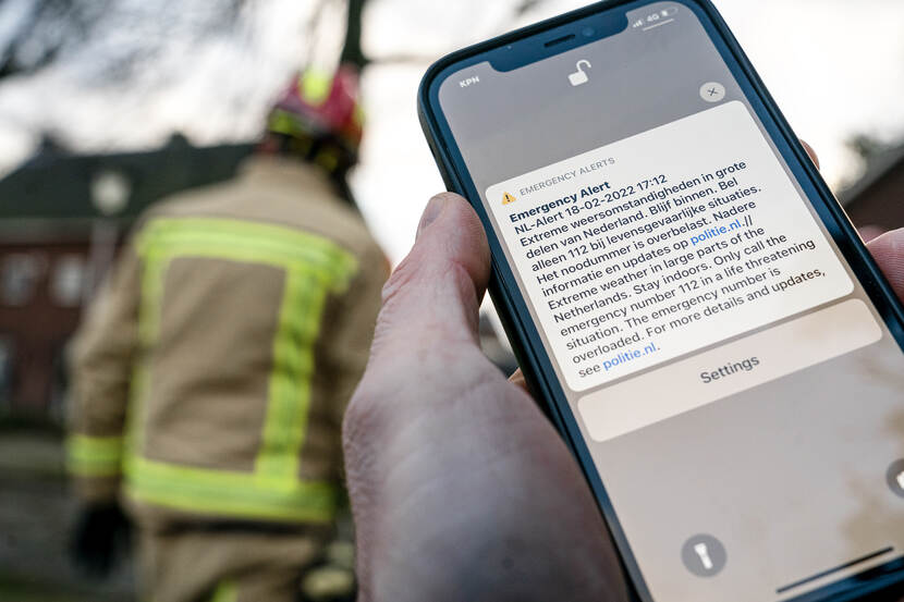 Close-up van een NL-Alert melding op een telefoon. Op de achtergrond staat een brandweerman en een huis.