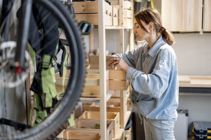 Persoon plaatst items in een houten kistje die op een houten stelling staan. Links hangt een fiets aan de muur.