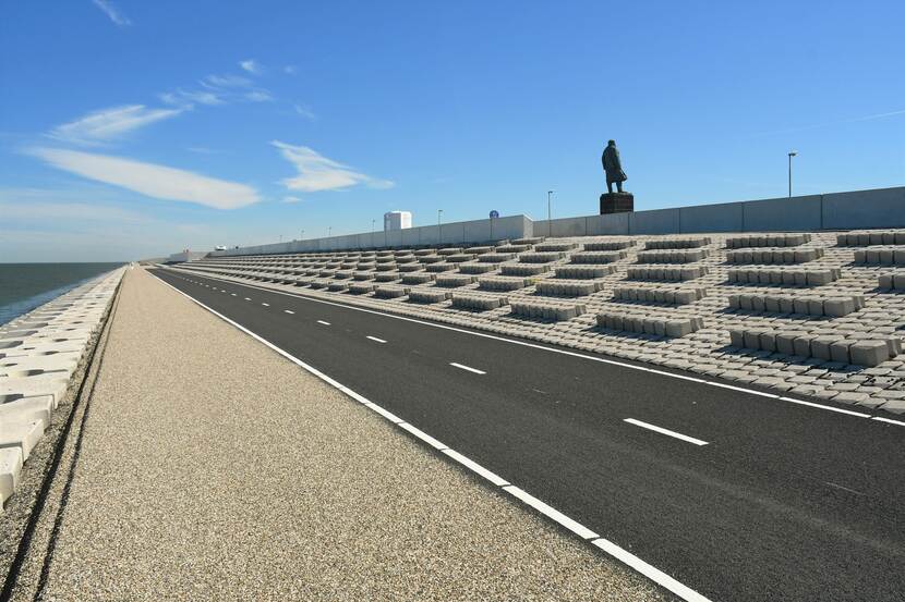 Beeld van een deel afsluitdijk. Rechts boven staat het beeld van ingenieur Cornelis Lely. Links is het water te zien met in het midden het fietspad op de afsluitdijk.