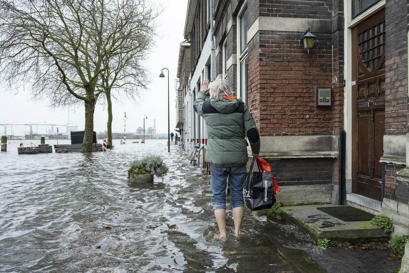 Een man loopt op blote voeten en met opgestroopte broekspijpen door een overstroomde straat. Hij zwaait naar buren die op een droge stoep staan. In zijn hand heeft hij een tas met spullen.