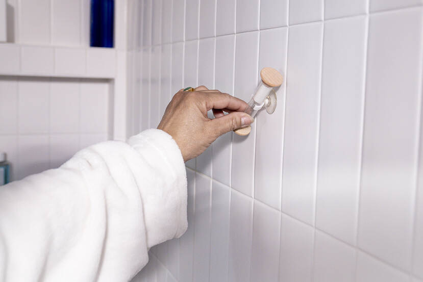 Een hand in een witte badjas draait een zandloper-douchetimer om, bevestigd met een zuignap aan een witte badkamerwand.
