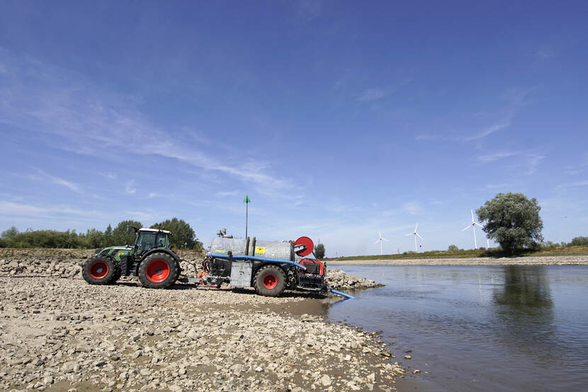 Een trekker met een watertank pompt water uit een rivier in een droge omgeving.