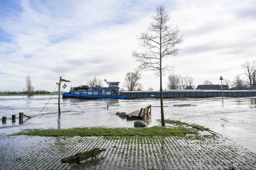 Een overstroomd grasveld met een scheefgezakte picknickbank en speeltoestel. Op de achtergrond vaart een binnenvaartschip over de rivier.